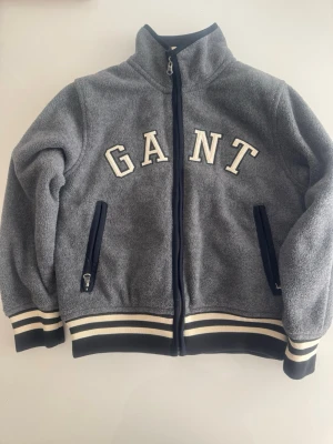 Grå barn fleecejacka med dragkedja GANT - Grå fleecejacka med hög krage och hel dragkedja framtill. Jackan har broderad GANT-logga på bröstet, två fickor med dragkedja och ribbade muddar samt nederkant med vita och marinblå ränder. Perfekt för en sportig och avslappnad stil.