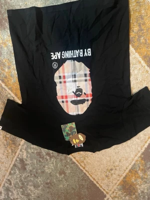 Svart BAPE t-shirt med rutigt tryck - Säljer en svart t-shirt från BAPE med det ikoniska apansiktet i ett rutigt mönster på bröstet och texten 'BY BATHING APE' ovanför. T-shirten har rund hals och korta ärmar. Perfekt för dig som gillar streetwear och vill sticka ut.
