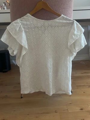Vit blus med volangärmar - Säljer en söt vit blus med strukturmönster och volangdetaljer på ärmarna. Blusen har en lös passform och är perfekt för dig som gillar en feminin och luftig stil. Materialet känns mjukt och har ett diskret rutmönster.