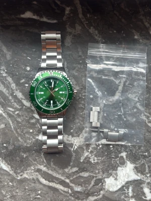 Grön sportig klocka i metall - Snygg klocka med grön urtavla och matchande grön bezel. Armbandet är i silverfärgad metall och har en klassisk länkdesign. Klockan har flera små urtavlor och tydliga siffror på bezeln. Perfekt accessoar för dig som gillar sportig stil.