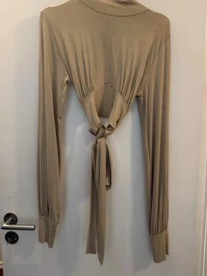 Beige långärmad topp med knyt - Beige långärmad topp med hög hals. Toppen har en öppen rygg med knytning 