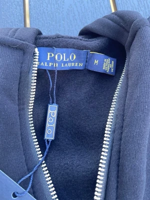 Marinblå hoodie från Polo Ralph Lauren - Snygg marinblå hoodie från Polo Ralph Lauren i storlek M. Hoodien har dragkedja framtill, huva och ribbade muddar. Tillverkad i mjukt bomullsmaterial som känns skönt mot huden. Klassisk och stilren design med diskreta detaljer och Polo-logga.