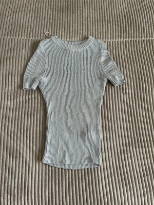 Ljusgrå ribbad topp med kort ärm - Säljer en ljusgrå ribbad topp med korta ärmar och rund hals. Toppen har en tight passform och är tillverkad i ett mjukt, stretchigt material som sitter snyggt på kroppen. Perfekt att styla med jeans eller kjol för en clean look. Tröjan är stretchig, alltså inte lika liten som på bilderna
