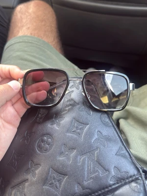 Svarta solglasögon från Prada - Snygga svarta solglasögon från Prada med fyrkantiga bågar och mörka glas. Bågarna har en stilren design med silvriga detaljer vid gångjärnen och tydlig Prada-logga på sidan. Perfekta för dig som vill ha en klassisk men trendig look. Prister kan diskuteras.