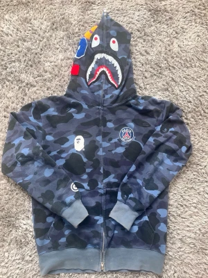 Blå camo hoodie från BAPE x PSG - Säljer en blå camo zip hoodie från BAPE x PSG med ikonisk hajdesign på huvan och broderade detaljer. Hoodien har PSG-logga på bröstet, BAPE-apan på sidan och färgglada applikationer på huvan. Tillverkad i mjuk bomullsblandning och har fickor fram.