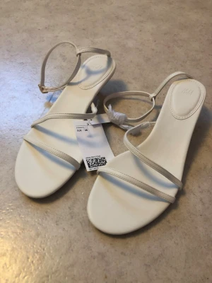 Vita sandaletter med smala remmar - Säljer ett par stilrena vita sandaletter med smala remmar och justerbart spänne runt ankeln. Sandalerna har en låg klack och en minimalistisk design som passar till många olika outfits. Materialet ser ut att vara konstläder.