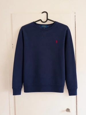 Polo Ralph Lauren sweatshirt - Polo Ralph Lauren sweatshirt i klassisk marinblå färg med röd broderad logga på bröstet 💙❤️ Storlek: M (10–12 år) • Mjuk och bekväm sweatshirt • Rund hals och ribbade muddar • Klassisk Polo Ralph Lauren-design • Fint begagnat skick utan defekter! Snygg och tidlös tröja som passar perfekt till vardags 👕✨