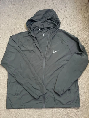 Svart Nike vindjacka med huva - Snygg svart vindjacka från Nike med dragkedja framtill och justerbar huva. Jackan har två sidofickor med dragkedja och en vit Nike-logga på bröstet. Tillverkad i lätt och tåligt material, perfekt för blåsiga dagar.