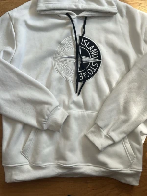 Vit hoodie från Stone Island - Säljer en vit hoodie från Stone Island med stor broderad logga i vitt och svart på bröstet. Hoodien har huva med svarta dragsnören och en klassisk magficka. Tillverkad i mjukt bomullsmaterial och har ribbade muddar vid ärmar och nederkant.