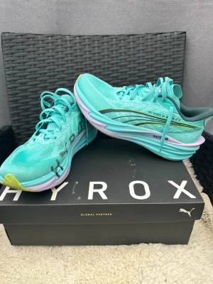 Turkosa Puma HYROX sneakers - Säljer ett par snygga turkosa Puma HYROX sneakers med mesh-överdel, gröna detaljer och vit/lila sula. Skorna har klassisk låg profil och sportig design med HYROX-tryck på sidan. Perfekta för dig som gillar att sticka ut med färg på fötterna.