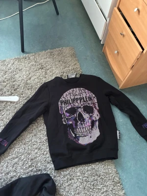 Svart Philipp Plein tröja med döskalle - Säljer en svart sweatshirt från Philipp Plein med stor döskalle i lila och silver på bröstet. Tröjan har rund hals, långa ärmar och coola detaljer med märkesnamnet på både bröstet och ena ärmen. Perfekt för dig som gillar edgy streetwear.