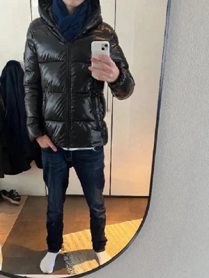 Svart glansig pufferjacka med huva - Säljer en svart, glansig pufferjacka med huva och dragkedja framtill. Jackan är tillverkad i ett djurfritt material och har en klassisk quiltad design. Perfekt för kalla vinterdagar och riktigt snygg till jeans eller andra byxor.