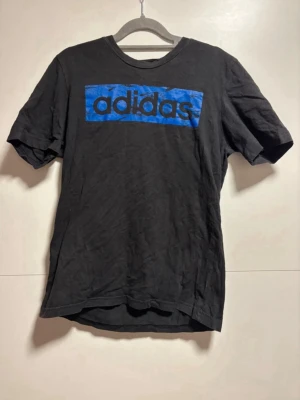 Svart Adidas t-shirt med blå logga - Svart t-shirt från Adidas med en stor blå logga på bröstet. Klassisk rund halsringning och korta ärmar. Tillverkad i mjuk bomull som känns skön mot huden. Perfekt för en avslappnad och sportig stil. Storlek S
