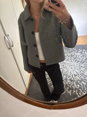 Grå ulljacka med knappar - Stilren grå jacka i ullmix med klassisk krage och stora svarta knappar framtill. Jackan har en rak passform. Perfekt för dig som gillar minimalistisk och tidlös stil. Använd en gång, nyskick (prickarna man ser är inte på jackan utan från spegeln)