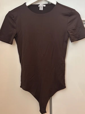 Brun kortärmad body  - Snygg brun body med korta ärmar och rund halsringning. Tillverkad i ett stretchigt och mjukt material som sitter tight mot kroppen. Perfekt att styla med jeans eller kjol för en clean look.