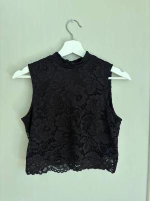 Svart spetsig croppad topp från Even&Odd - Säljer en svart croppad topp från Even&Odd med blommig spets och hög hals. Toppen är ärmlös och har ett elegant, genombrutet mönster som ger en trendig look. Perfekt att styla med höga jeans eller kjol.