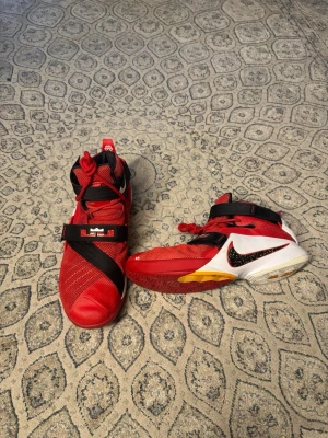 Nike LeBron James Soldier 9 University - Snygga Nike LeBron sneakers i rött med svarta och vita detaljer samt gula inslag på sulan. Skorna har högt skaft, mesh- och syntetmaterial, samt kardborreband över vristen för extra support. Ikonisk LeBron-logga på remmen och klassisk Nike Swoosh på sidan. Nike LeBron James Soldier 9 University, är inte så jätte slitna, storlek 48,5 EU, skickar samma dag eller dagen efter :)