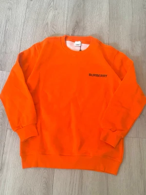 Orange sweatshirt från Burberry - Säljer en riktigt snygg orange sweatshirt från Burberry med svart logga på bröstet. Tröjan har rund halsringning, långa ärmar och ribbade muddar vid ärmslut och nederkant. Perfekt för dig som vill sticka ut med färg! Materialet känns mjukt och skönt.