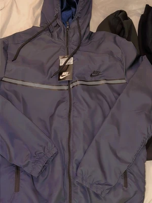 Blå Nike vindjacka med huva - Snygg blå vindjacka från Nike med huva och hel dragkedja framtill. Jackan har svarta detaljer, reflekterande rand över bröstet och elastiska muddar vid ärmslut. Perfekt för blåsiga dagar och sportiga outfits. Helt ny st Medium 