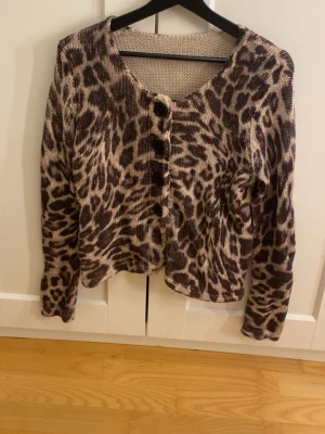 Leopardmönstrad kofta med knappar - Snygg leopardmönstrad kofta i beige och brunt med långa ärmar och rund halsringning. Koftan har stora svarta knappar framtill och är tillverkad i ett mjukt stickat material. Perfekt för dig som vill sticka ut lite extra.