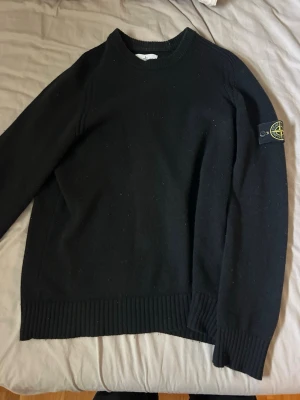 Svart stickad tröja från Stone Island - Snygg svart stickad tröja från Stone Island med klassisk patch på vänster ärm. Tröjan har rund halsringning, ribbade muddar och är tillverkad i mjukt material som passar perfekt till höst och vinter.