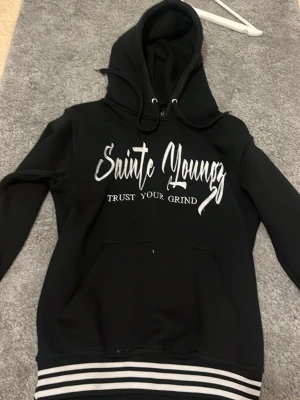 Svart hoodie med text och ränder - Svart hoodie med vit text 'Sainte Youngz Trust Your Grind' på framsidan och citat på ryggen. Huva med dragsko och magficka. Ribbstickade muddar och nederkant med vita och svarta ränder. Perfekt för en chill och street-inspirerad look.