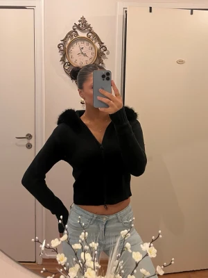 Svart hoodie med fuskpäls på huvan - Snygg svart zip hoodie med croppad passform och fluffig fuskpäls runt huvan. Tröjan har långa ärmar och dragkedja framtill, perfekt för en trendig och mysig look. Passar till både jeans och kjol för en cool vardagsstil.