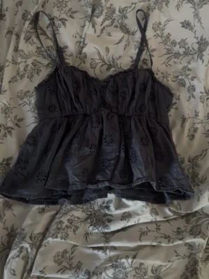 Mörkgrå broderad linnetopp Hollister - Söt mörkgrå linnetopp från Hollister med tunna axelband och broderade blommönster. Toppen har en luftig passform och smockdetalj upptill för extra komfort. Perfekt för varma dagar och enkel att styla med jeans eller kjol.