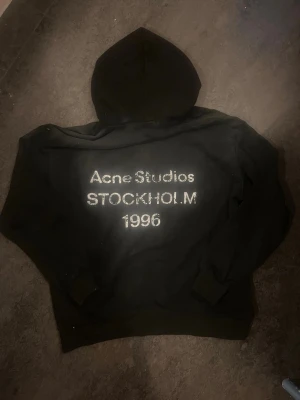 Svart hoodie Acne Studios Stockholm 1996 - Svart hoodie från Acne Studios med stor huva och tryck på ryggen där det står 'Acne Studios STOCKHOLM 1996' i vitt. Klassisk passform med ribbade muddar vid ärmar och midja. Perfekt för en avslappnad och trendig streetwear-look. Kvitto finns