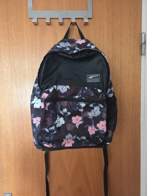 Svart ryggsäck med blommigt mönster - Snygg svart ryggsäck med blommigt mönster i rosa, vitt och lila. Ryggsäcken har ett stort huvudfack, en framficka med dragkedja och nätficka på sidan. Justerbara axelband med blommiga detaljer. Perfekt för skolan eller stan.