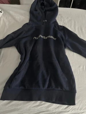 Mörkblå hoodie Peak Performance - Snygg mörkblå hoodie från Peak Performance med stor huva och magficka. Klassisk logga broderad på bröstet. Tillverkad i mjukt bomullsmaterial som känns skönt mot huden. Perfekt för en avslappnad och sportig stil. Har en fläck på ryggen som jag är osäker på går bort eller inte men det märks inte så mycket.