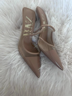 Beige transparenta pumps från Zara - Säljer ett par snygga beige pumps från Zara med transparent plastdetalj och spetsig tå. Skorna har en låg klack och en tunn rem över foten för extra stil. Perfekta för dig som vill ha en trendig och modern look.