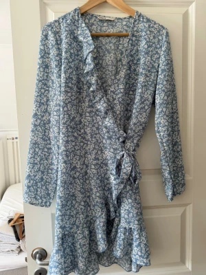 Blommig blå omlottklänning med volang - Söt blå kortklänning med vitt blommönster och volangdetaljer. Omlottmodell med knyt i midjan och långa ärmar. Perfekt för vår och sommar när du vill känna dig extra fin.