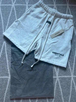 Grå shorts från Essentials Fear of God - Snygga ljusgrå shorts från Essentials Fear of God med elastisk midja och långa snören framtill. Shortsen har en relaxed passform och logga tryckt på vänster ben. Perfekta för chill dagar och enkel stil.