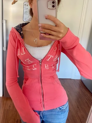Korall rosa tajt Hollister zip hoodie med broderi - Gullig korallrosa zip hoodie med vit broderad hollister text över bröstet, strl M omsydd till xxs! Följ för mer liknande hollister och abercrombie zipups