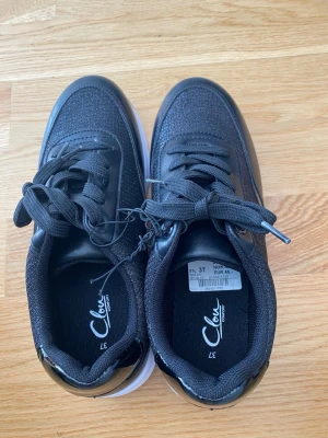 Svarta sneakers från Clou - Snygga svarta sneakers från Clou med textil och syntetmaterial. Skorna har klassisk låg profil, svarta skosnören och en vit sula som kontrast. Perfekta för dig som gillar stilrena och bekväma sneakers till vardagslooken.