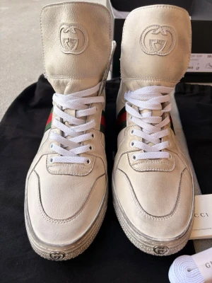 Beige höga sneakers från Gucci - Snygga höga sneakers från Gucci i beige läder med vita snören och klassiska gröna och röda ränder på sidan. Tungan har präglad Gucci-logga och sulan har diskret logodetalj framtill. Perfekta för dig som vill ha en clean och exklusiv look.