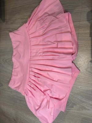 Rosa plisserad skort - Supersöt rosa skort med plisserad kjoldel och inbyggda shorts. Tillverkad i ett mjukt och stretchigt material som ger en bekväm passform. Perfekt för dig som vill ha kjol-looken men ändå känna dig bekväm och fri att röra dig. Den är storlek M men gansk kort. Oanvänd