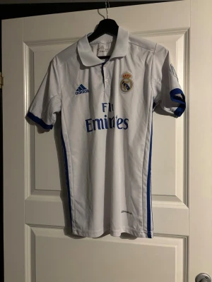 Real Madrid Ronaldo matchtröja vintage - Vit Real Madrid matchtröja från Adidas med blå detaljer, klubbmärke och sponsortryck på framsidan. Nummer 7 och Ronaldo på ryggen. Klassisk pikékrage och korta ärmar, perfekt för fotboll eller som supporterplagg. Köpt på vintage butik