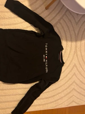 Svart långärmad tröja Tommy Hilfiger - Svart långärmad tröja från Tommy Hilfiger med logga och texten 'TOMMY HILFIGER EST. 1985' på bröstet. Tröjan har rund halsringning med en snygg detalj i rött, vitt och blått på insidan av kragen. Perfekt för en stilren och avslappnad look.