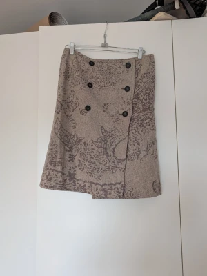 Beige kjol med knappar och mönster - Snygg beige kjol med dekorativa mörka knappar framtill och ett diskret, paisley-liknande mönster i brunt. Kjolen har omlott-look och är lätt utställd. Perfekt för dig som gillar unika detaljer och stilren design.