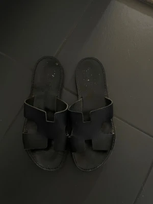 Handgjorda svart läder tofflor från Grekland  - Säljer ett par svarta slip-in sandaler med breda remmar i läder och öppen tå. Sandalerna har en stilren design med ett H-format mönster på ovandelen och platt sula. Perfekta för sommaren och enkla att matcha till olika outfits.