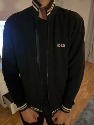 Svart bomberjacka från BOSS - Säljer en svart bomberjacka från BOSS med vita och beige ränder på krage, ärmslut och nederkant. Jackan har dragkedja framtill och BOSS-logga broderad på bröstet. Klassisk och stilren modell som passar perfekt till streetwear. Dock fungerar ej kedjan, därav priset