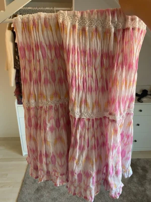 Långkjol från Gina Tricot i rosa mönster - Säljer en flowy långkjol från Gina Tricot med vitt, rosa och gult mönster. Kjolen har volangdetaljer och spets längs sömmarna, samt ett lätt och luftigt tyg som passar perfekt till varmare dagar.