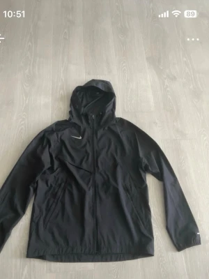 Svart Nike vindjacka med huva - Snygg svart vindjacka från Nike med hel dragkedja framtill och praktisk huva. Jackan har två sidofickor och diskret Nike-logga på bröstet. Tillverkad i lätt och vindtätt material, perfekt för blåsiga dagar.