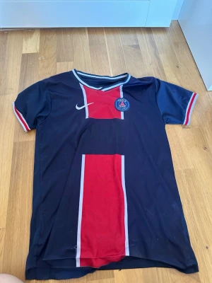PSG matchtröja i mörkblått och rött - Snygg PSG matchtröja från Nike i mörkblått med röd vertikal rand och vita detaljer. Tröjan har korta ärmar, klubbmärke på bröstet och klassisk sportig design. Perfekt för fotbollsträning eller att bära till vardags.