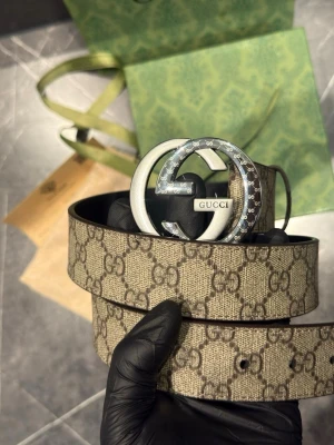 Gucci GG Supreme beige bälte - Säljer ett klassiskt Gucci GG Supreme bälte i beige och brunt med det ikoniska GG-mönstret. Bältet har ett stort, silverfärgat GG-spänne i metall och är tillverkat i canvas med läderdetaljer. Perfekt för att lyfta vilken outfit som helst.