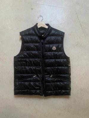 Svart dunväst från Moncler - Snygg svart dunväst från Moncler med quiltad design och hög krage. Västen har två dragkedjeförsedda fickor framtill och Moncler-logga på bröstet. Perfekt för lager-på-lager och ger en clean look till din outfit. Saknar helt skador. Storlek 4 = M. Självklart äkta.
