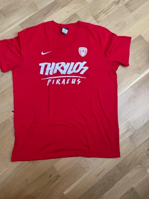 Röd Nike t-shirt Thrylos Piraeus - Röd t-shirt från Nike med trycket 'Thrylos Piraeus' och klubbmärke på bröstet. Klassisk passform med rund hals och korta ärmar. Perfekt för dig som gillar sportig stil och vill sticka ut med en färgstark tröja.