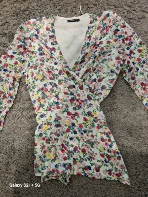 Blommig omlottklänning med långa ärmar - Söt omlottklänning med vit bas och färgglatt blommönster i rosa, blått, gult och grönt. Klänningen har långa ärmar och en djup v-ringning. Materialet ser lätt och luftigt ut, perfekt för vår och sommar.